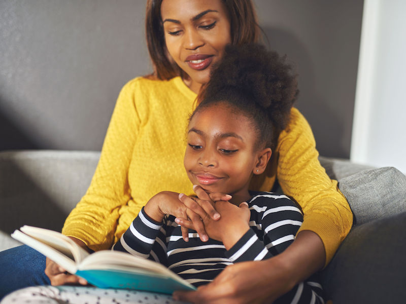 Black mom and daughter reading a book sitting on sofa smiling