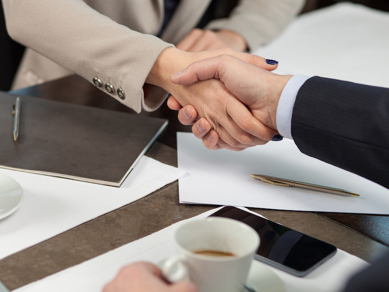 We made a deal. Close up of arms of successful two business partners shaking hands. They are sitting at the table near documents in cafe. The businessman and woman are drinking coffee