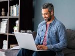 Successful mature indian businessman sitting on couch typing on laptop with wireless earphones. Mixed race businessman sitting on couch while working from home during video call. Happy multiethnic business man in casual clothing working on computer.